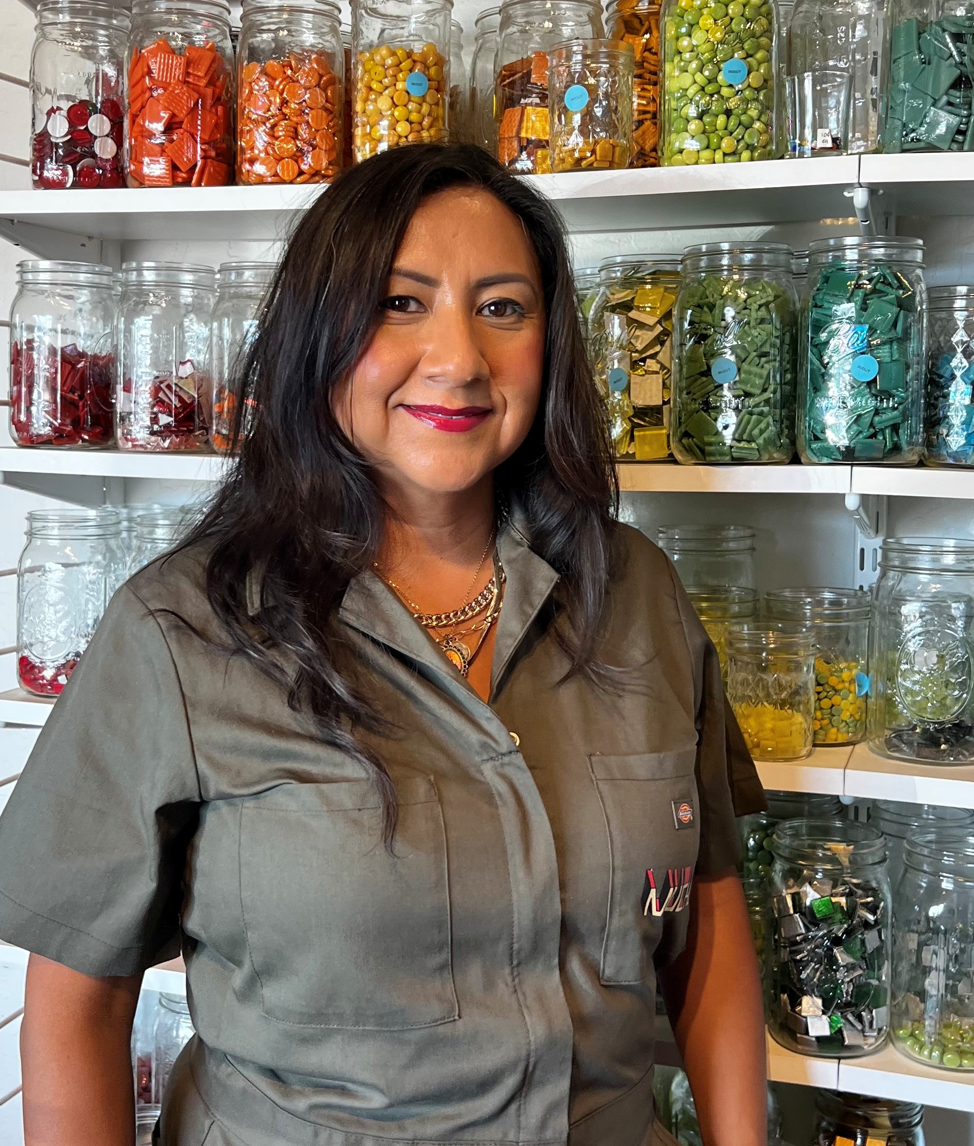 sandra A smiling woman with long brown hair and red lipstick stands in front of mosaic glass in jars